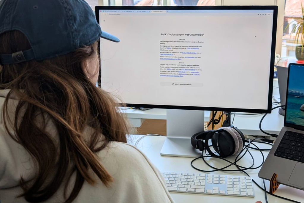 Person with long hair and a cap sits at a desk using a large monitor displaying a login screen; keyboard, headphones, and a laptop on the desk.