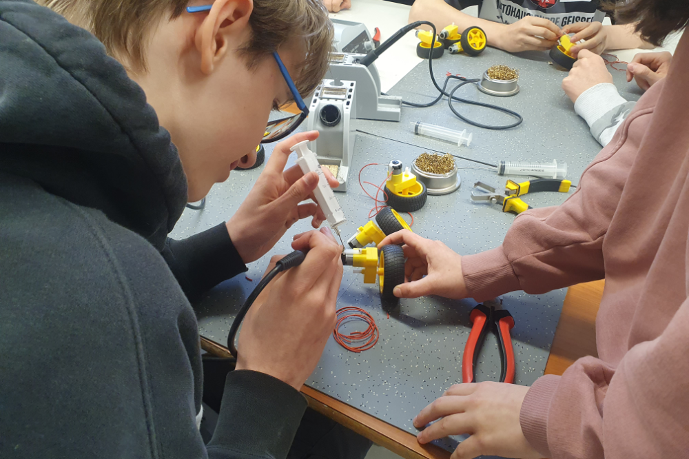 Students assembling small robotic wheels with tools and wires on a workbench.